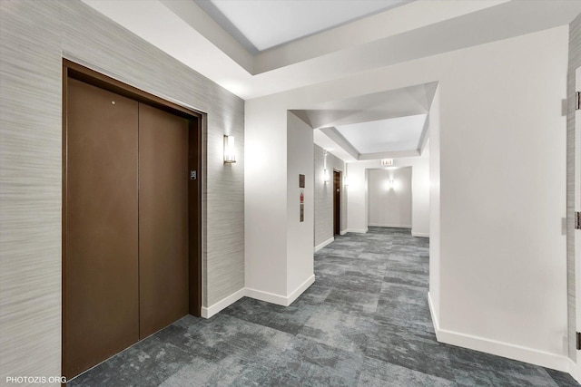 hall featuring a tray ceiling, elevator, baseboards, and dark colored carpet