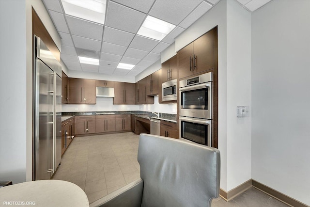 kitchen featuring baseboards, a drop ceiling, appliances with stainless steel finishes, under cabinet range hood, and dark countertops