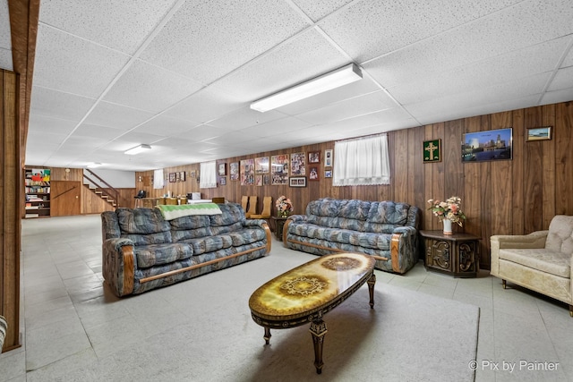 living area featuring stairs, a drop ceiling, and wooden walls