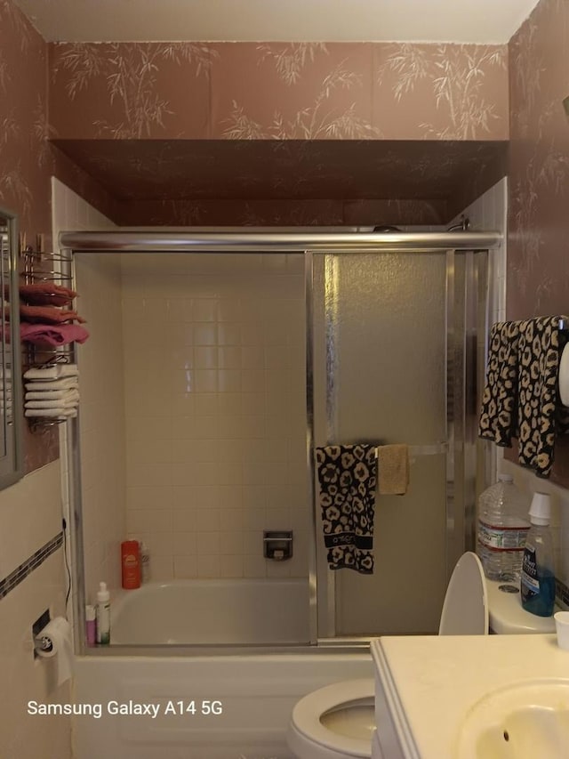 bathroom with combined bath / shower with glass door, vanity, toilet, and wallpapered walls