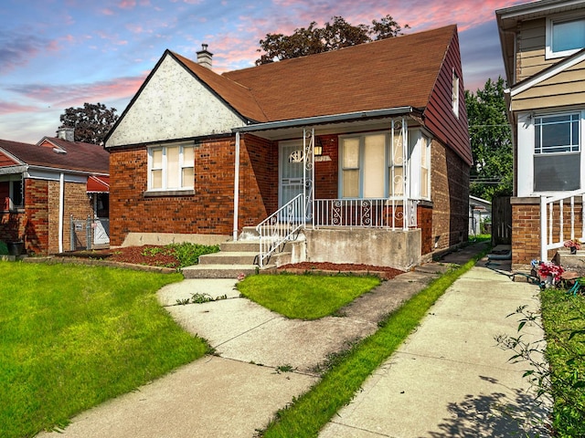 bungalow featuring covered porch, brick siding, roof with shingles, a front lawn, and a chimney