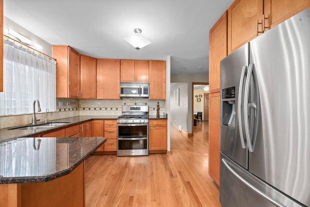 kitchen featuring light wood finished floors, backsplash, appliances with stainless steel finishes, a sink, and dark stone countertops