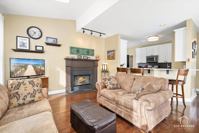 living area featuring dark wood finished floors, a glass covered fireplace, track lighting, and baseboards