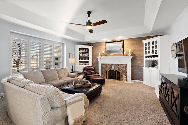 carpeted living area with a tray ceiling, a brick fireplace, wood walls, and ceiling fan