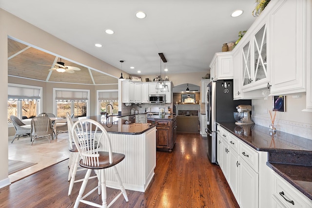 kitchen featuring a kitchen bar, an island with sink, a ceiling fan, dark countertops, and stainless steel appliances