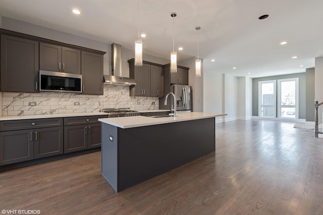 kitchen with a sink, light countertops, appliances with stainless steel finishes, wall chimney exhaust hood, and an island with sink