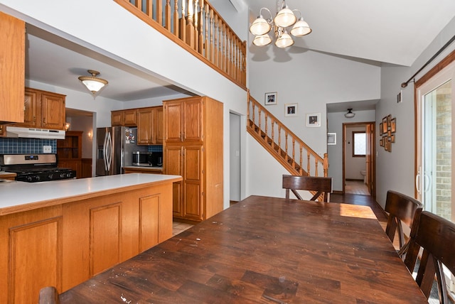 kitchen featuring tasteful backsplash, under cabinet range hood, brown cabinetry, a notable chandelier, and stainless steel appliances