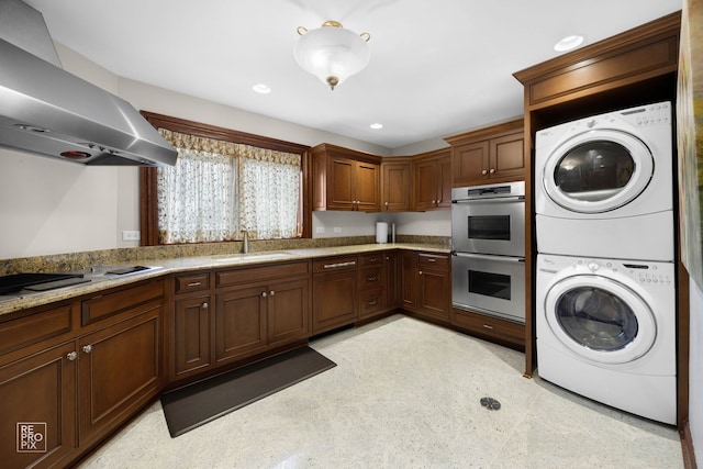 kitchen with stacked washer and dryer, stovetop with downdraft, stainless steel double oven, a sink, and wall chimney exhaust hood