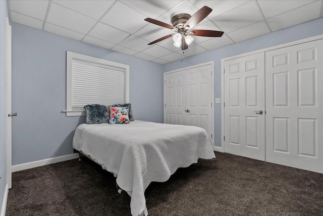 bedroom featuring two closets, baseboards, a drop ceiling, a ceiling fan, and dark colored carpet