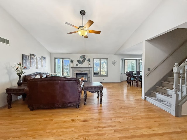 living area featuring visible vents, light wood-style floors, a ceiling fan, a glass covered fireplace, and stairs