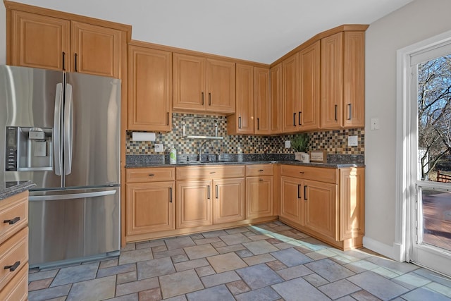 kitchen featuring a sink, tasteful backsplash, stone finish floor, dark stone countertops, and stainless steel fridge