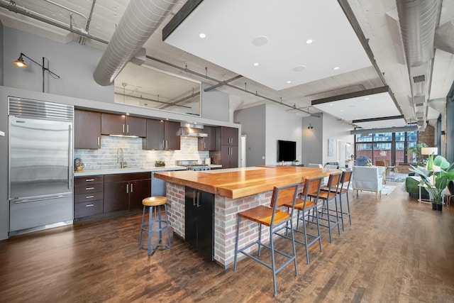 kitchen featuring dark wood-style floors, wooden counters, wall chimney range hood, and built in fridge