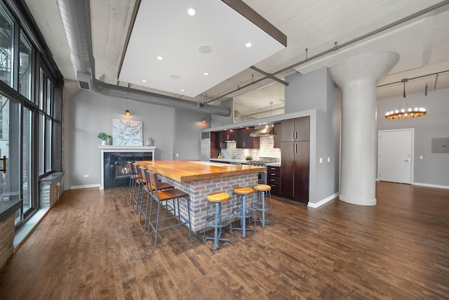 kitchen with dark wood-style flooring, a kitchen breakfast bar, a lit fireplace, wooden counters, and wall chimney range hood