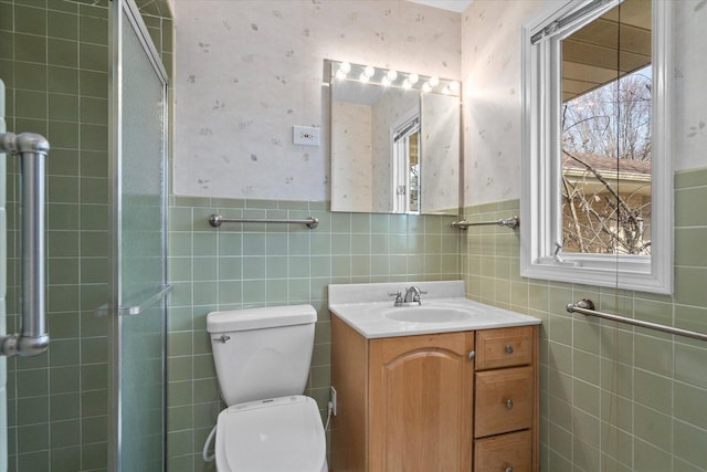 bathroom with wallpapered walls, toilet, a tile shower, wainscoting, and vanity