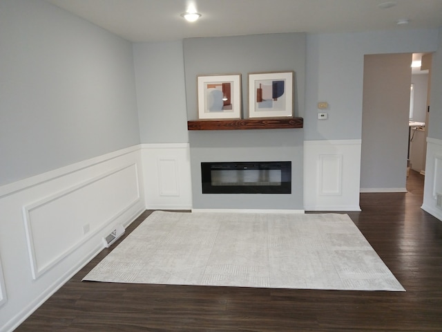 unfurnished living room featuring wainscoting, a decorative wall, dark wood-type flooring, and a glass covered fireplace