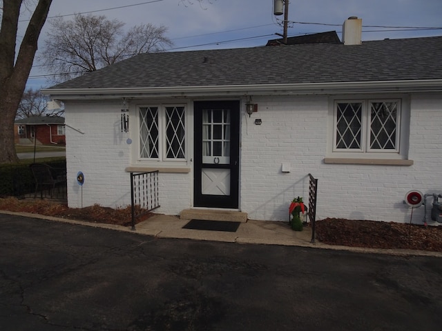 doorway to property featuring brick siding, a chimney, and a shingled roof