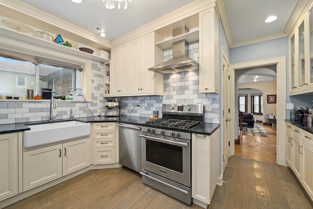 kitchen with open shelves, appliances with stainless steel finishes, arched walkways, wall chimney exhaust hood, and a sink