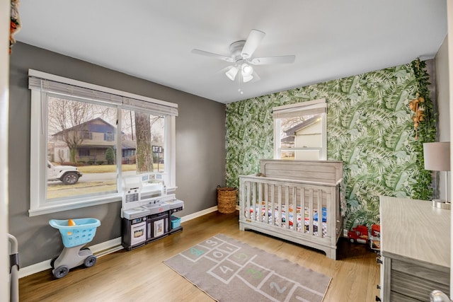 bedroom featuring wallpapered walls, baseboards, ceiling fan, wood finished floors, and a nursery area