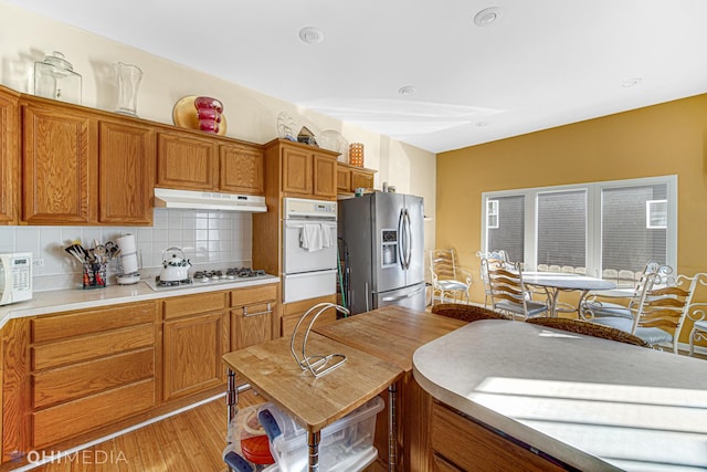 kitchen featuring under cabinet range hood, white appliances, light countertops, brown cabinets, and decorative backsplash