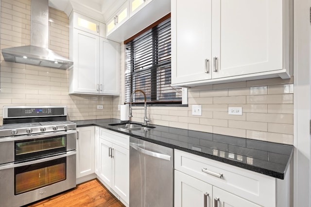 kitchen with stainless steel appliances, a sink, white cabinetry, wall chimney range hood, and decorative backsplash