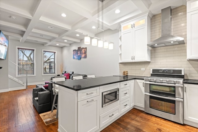 kitchen featuring coffered ceiling, dark countertops, wall chimney exhaust hood, appliances with stainless steel finishes, and a peninsula