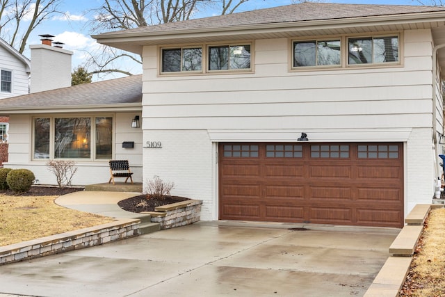 tri-level home with driveway, roof with shingles, a garage, brick siding, and a chimney