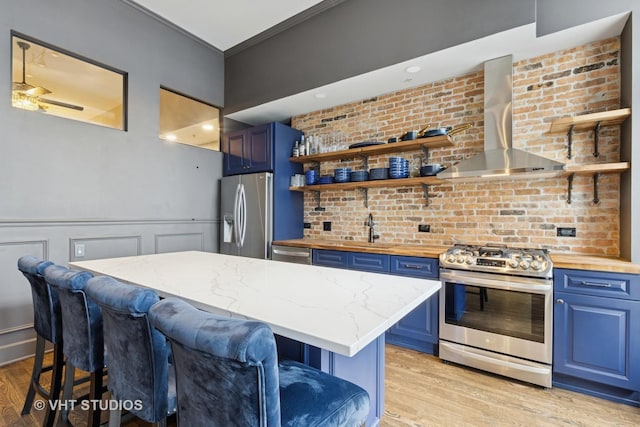 kitchen featuring blue cabinets, wood counters, appliances with stainless steel finishes, and wall chimney exhaust hood