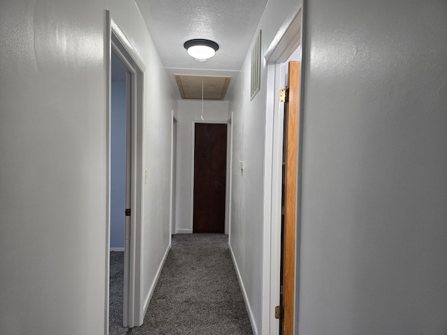 hallway featuring attic access, dark colored carpet, a textured ceiling, and baseboards