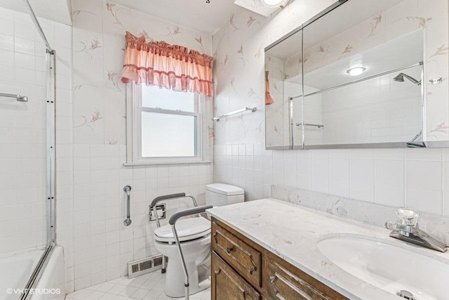 bathroom featuring visible vents, toilet, vanity, shower / bath combination with glass door, and tile walls