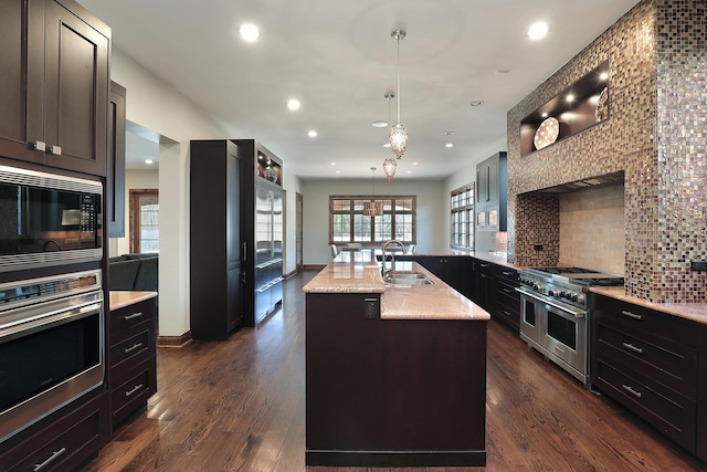 kitchen with dark wood finished floors, an island with sink, a sink, decorative backsplash, and appliances with stainless steel finishes