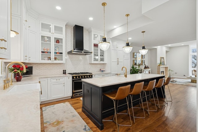 kitchen with wall chimney range hood, an island with sink, high end range, white cabinetry, and a sink