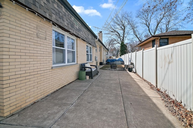 view of side of property featuring a patio, fence, a chimney, a shingled roof, and brick siding