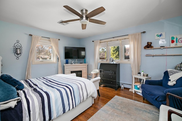 bedroom featuring visible vents, baseboard heating, wood finished floors, a glass covered fireplace, and a ceiling fan