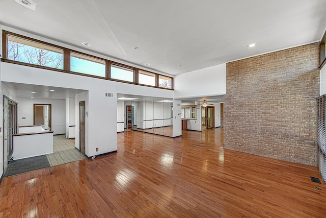 unfurnished living room with visible vents, a towering ceiling, brick wall, ceiling fan, and hardwood / wood-style floors