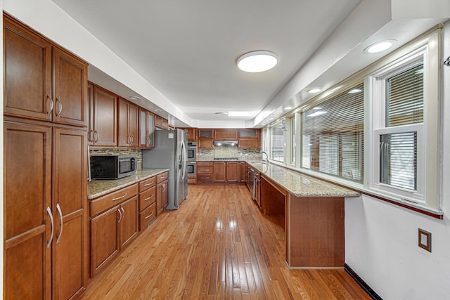 kitchen with brown cabinets, stainless steel appliances, decorative backsplash, light wood-type flooring, and under cabinet range hood