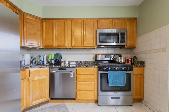 kitchen featuring brown cabinets, dark stone countertops, stainless steel appliances, tile walls, and light tile patterned floors