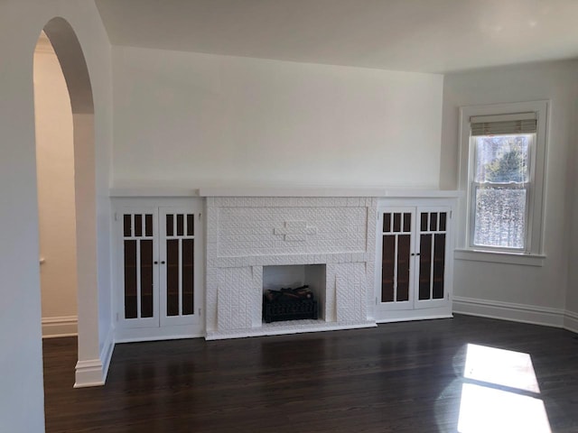 unfurnished living room with arched walkways, a brick fireplace, baseboards, and wood finished floors