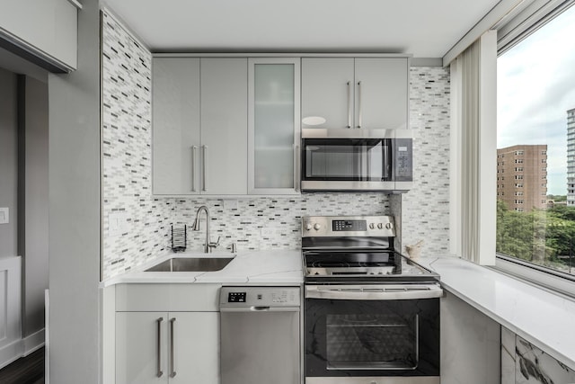 kitchen featuring gray cabinetry, a sink, backsplash, appliances with stainless steel finishes, and glass insert cabinets