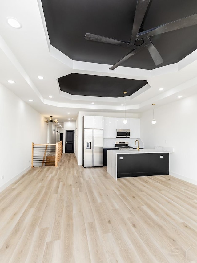 kitchen with a center island with sink, stainless steel appliances, a raised ceiling, light wood-style floors, and white cabinetry