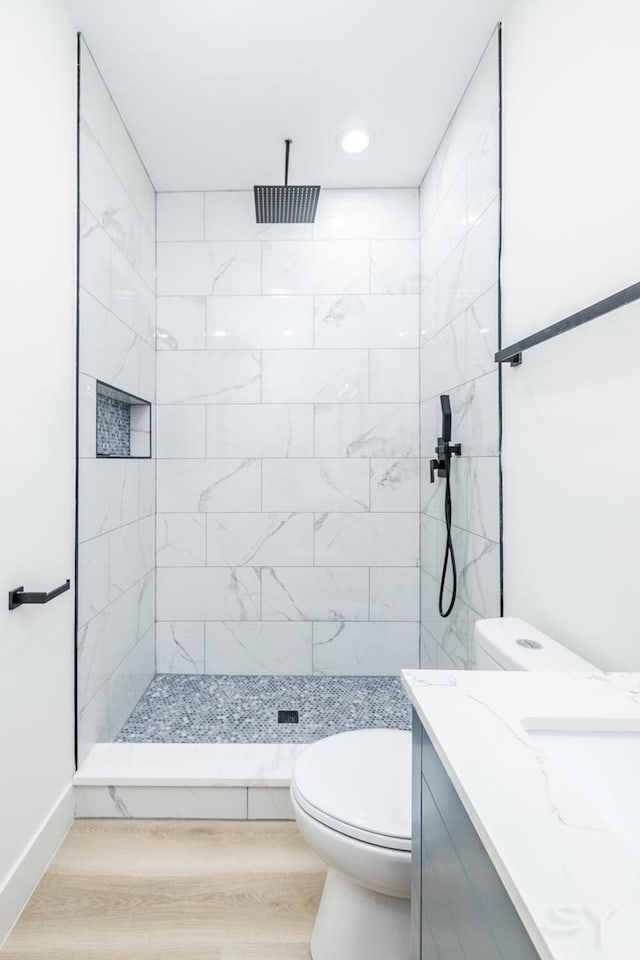 bathroom with toilet, a tile shower, vanity, wood finished floors, and baseboards