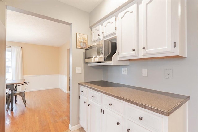 kitchen with light wood-style flooring, stainless steel microwave, dark countertops, white cabinets, and baseboards