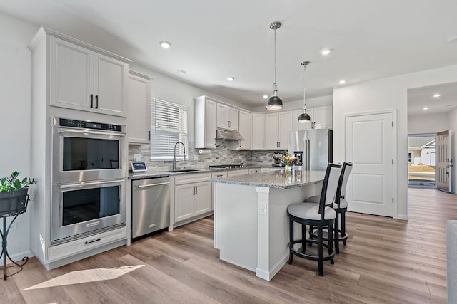 kitchen with a center island, under cabinet range hood, light stone counters, appliances with stainless steel finishes, and a sink