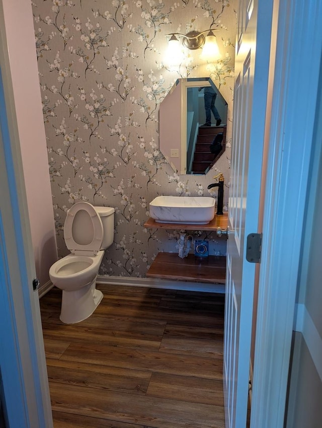 bathroom featuring toilet, wood finished floors, a sink, baseboards, and wallpapered walls