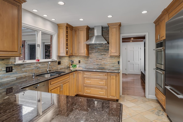 kitchen with backsplash, dark stone countertops, appliances with stainless steel finishes, wall chimney exhaust hood, and a sink