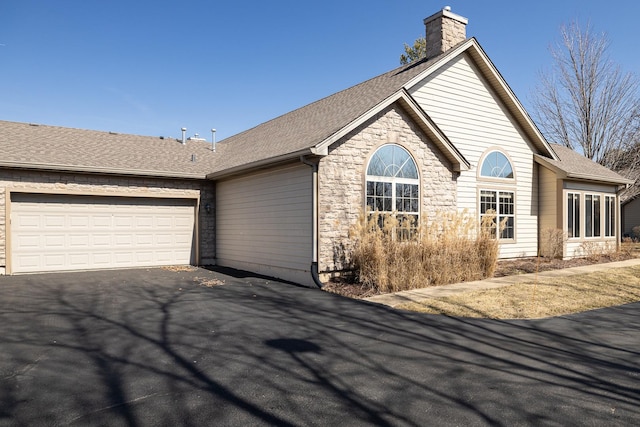 view of front facade featuring an attached garage, roof with shingles, a chimney, stone siding, and driveway