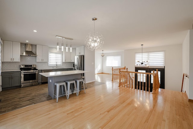 kitchen featuring an inviting chandelier, stainless steel appliances, decorative backsplash, light wood-style floors, and wall chimney exhaust hood