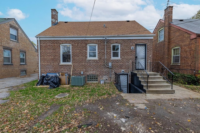 back of property with a shingled roof, a chimney, central AC unit, and brick siding