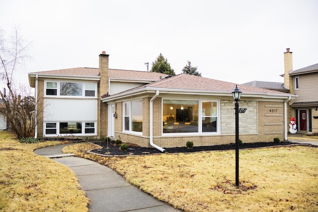 tri-level home with roof with shingles, brick siding, a chimney, and a front lawn