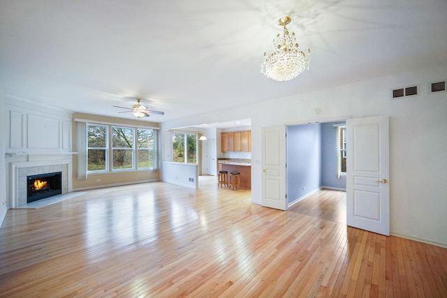 unfurnished living room featuring ceiling fan with notable chandelier, a fireplace, visible vents, baseboards, and light wood-type flooring