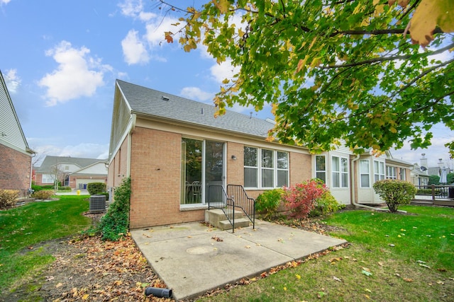 rear view of property featuring a shingled roof, a patio, a yard, central AC, and brick siding